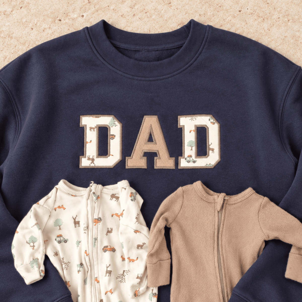 A navy crewneck sweatshirt laid flat on a light tan textured background, featuring the word 'DAD' in large varsity-style letters with woodland animal prints. Two baby onesies are placed in front of the sweatshirt: one cream zip-front romper with a woodland print and one solid tan zip-front romper.
