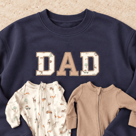 A navy crewneck sweatshirt laid flat on a light tan textured background, featuring the word 'DAD' in large varsity-style letters with woodland animal prints. Two baby onesies are placed in front of the sweatshirt: one cream zip-front romper with a woodland print and one solid tan zip-front romper.