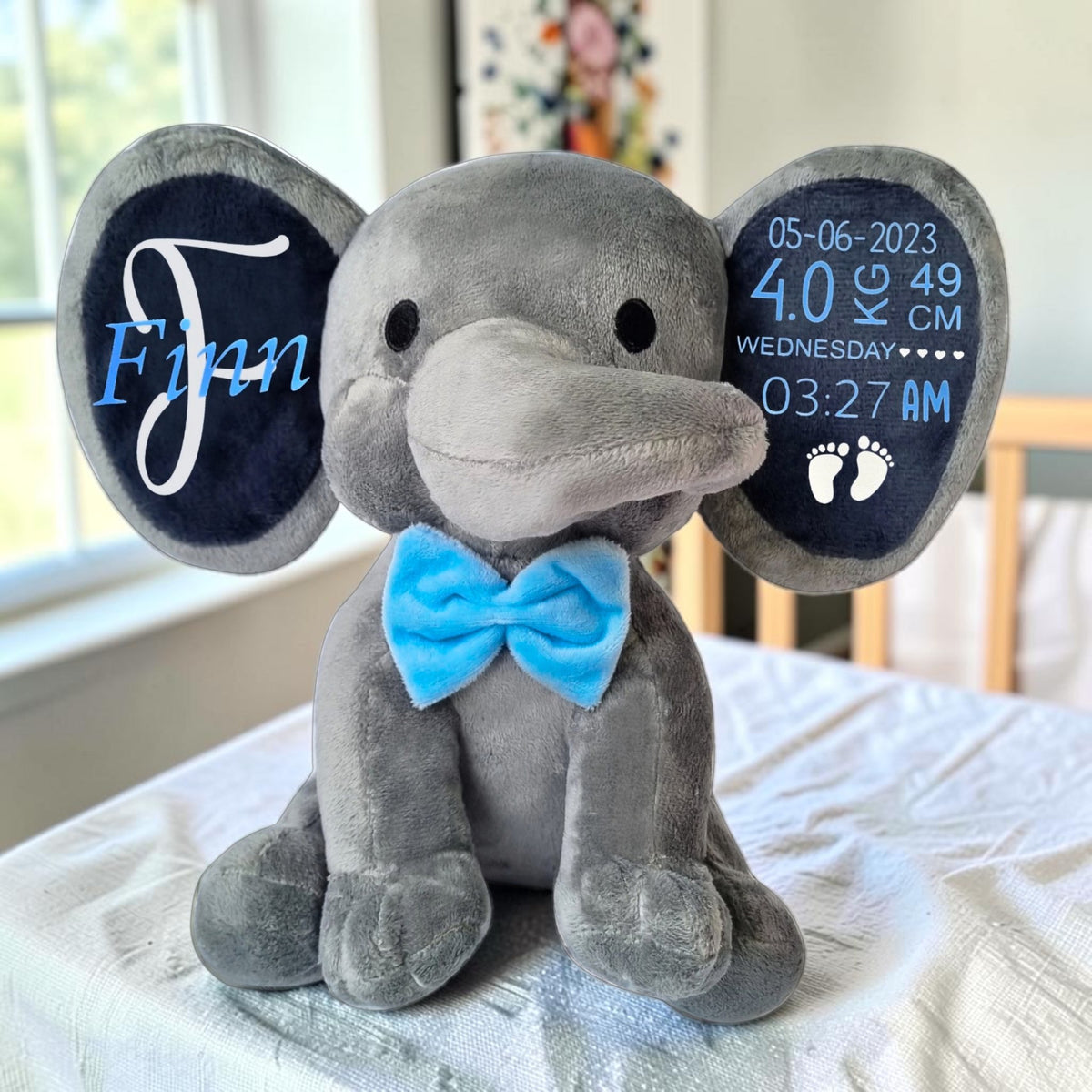 A personalized grey plush elephant .  The left ear has a blue background with the name 'Finn' in white script and light blue 'J'.