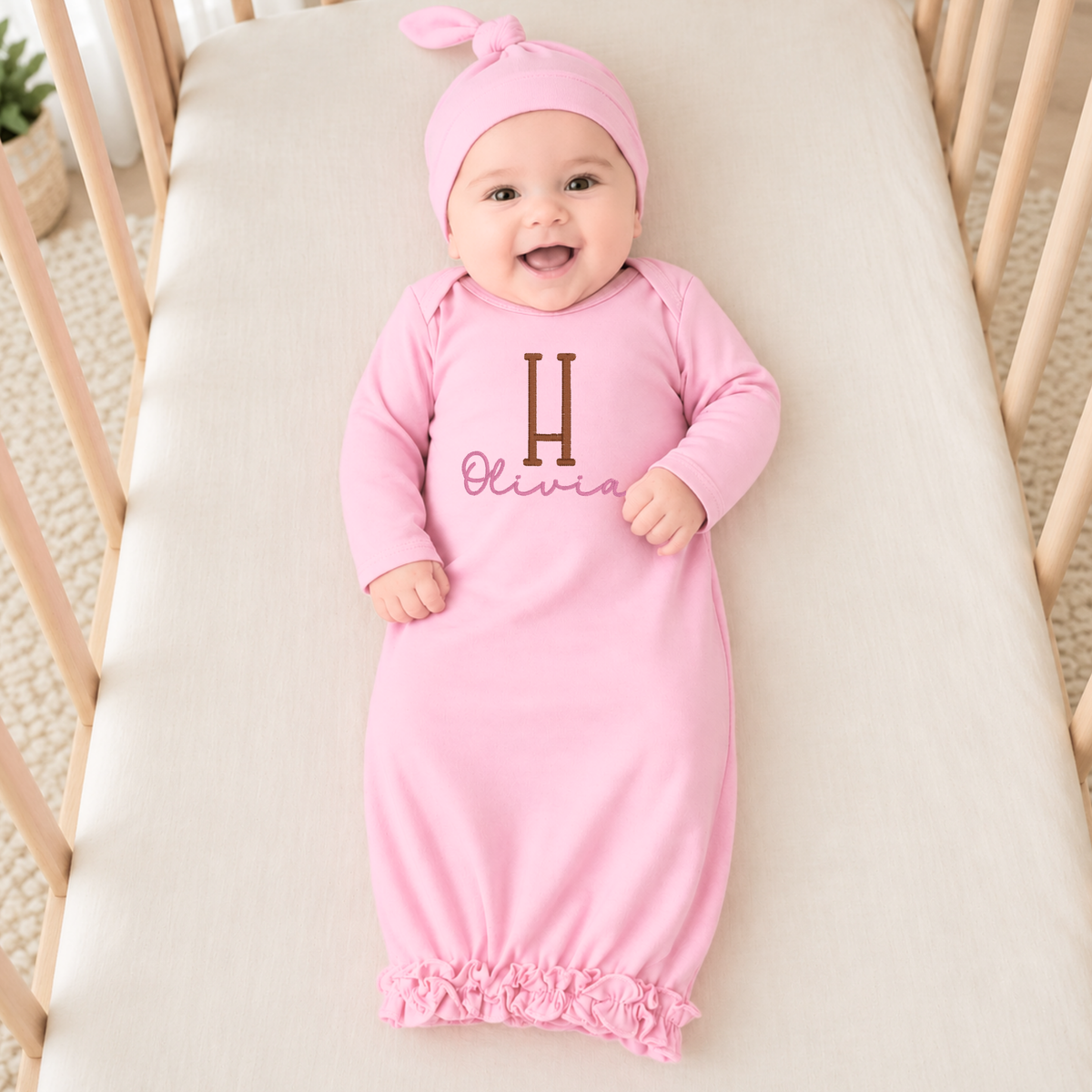 A baby wearing a pink long-sleeve sleep gown and matching pink bow headband, lying on a cream mattress in a wooden crib. The sleep sack has a brown embroidered letter 'H' and the name 'Olivia' in pink script.